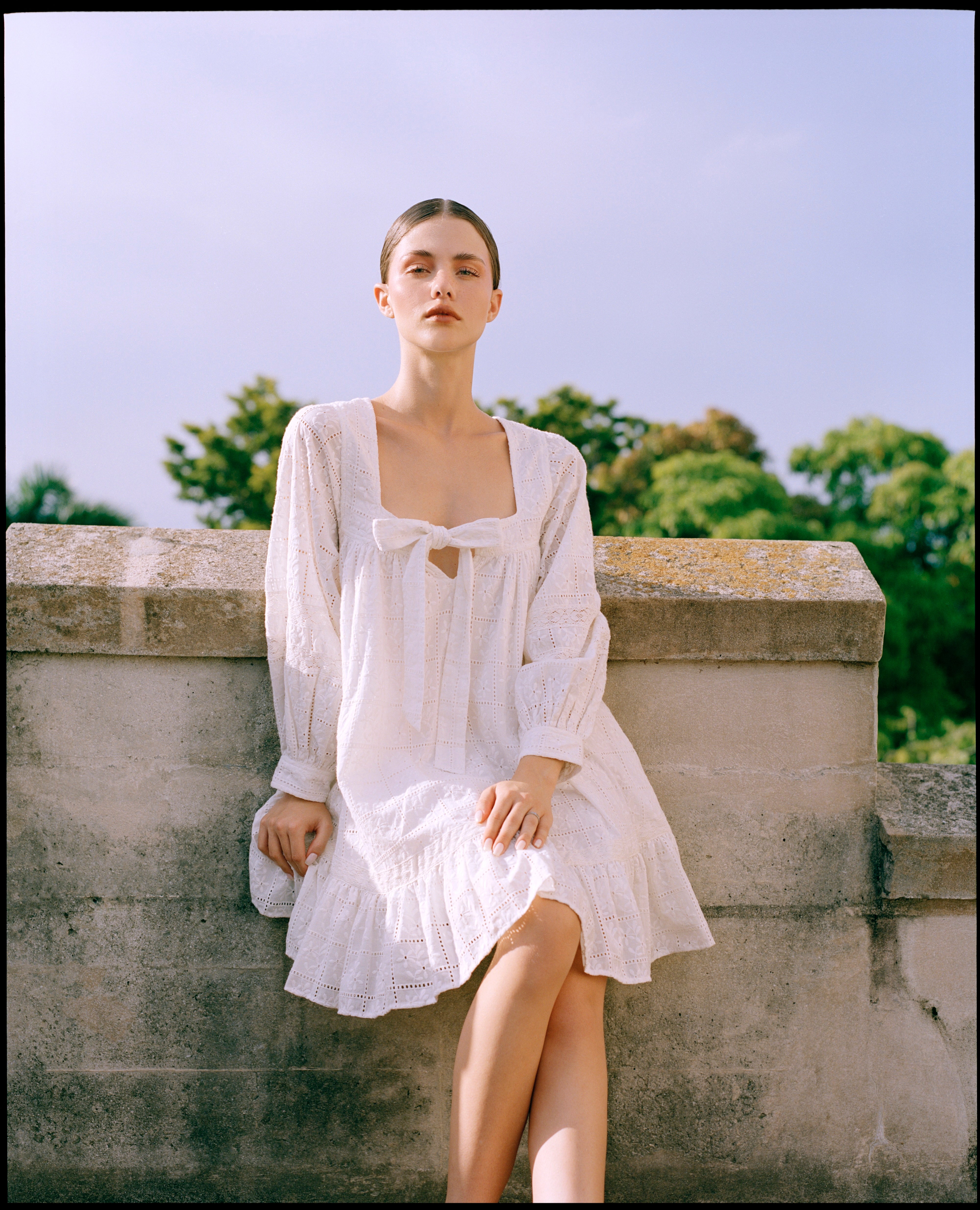 Woman in a white dress sitting on a stone wall with trees and blue sky in the background