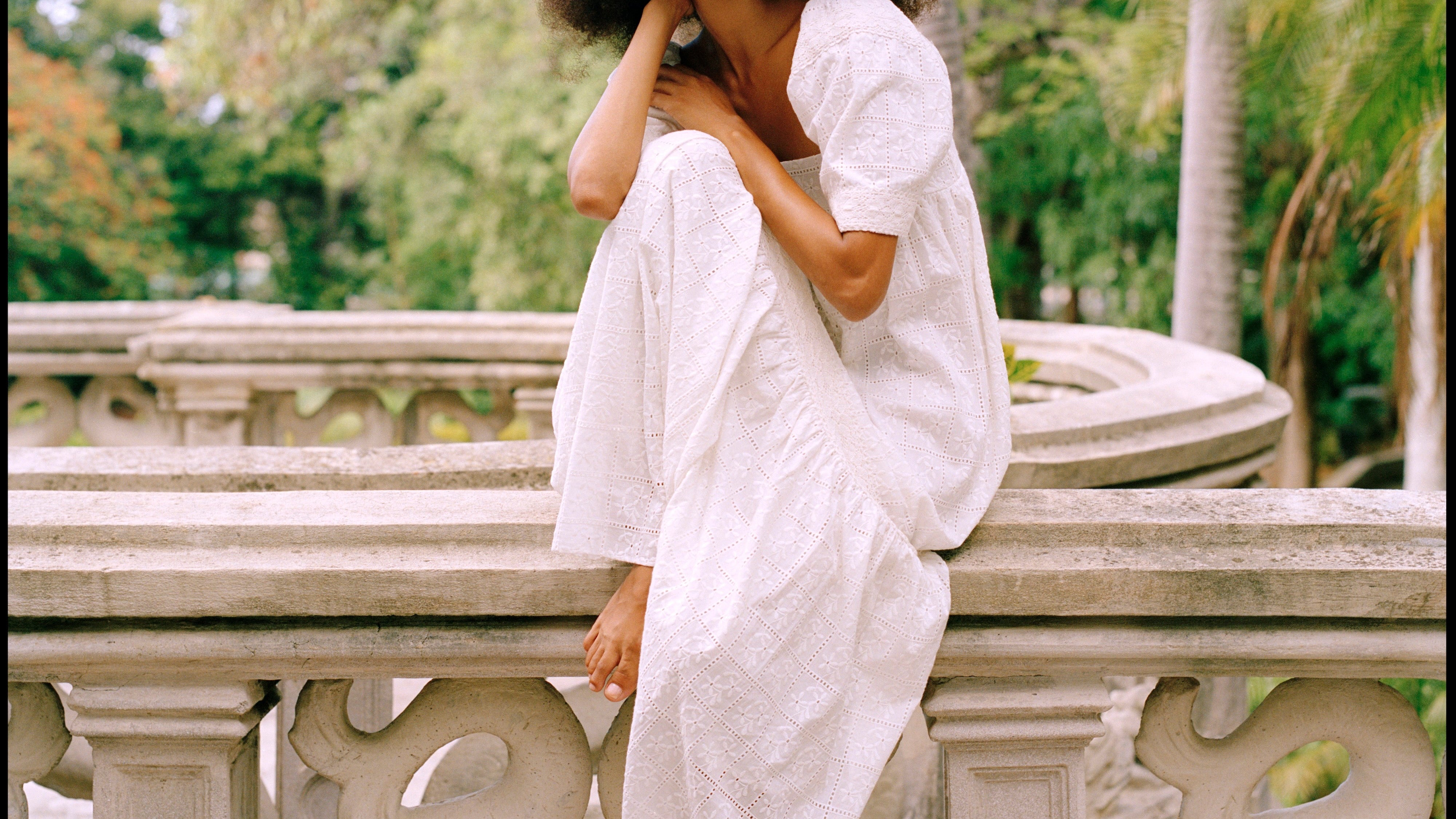 Woman in a white dress sitting on a stone railing with trees in the background