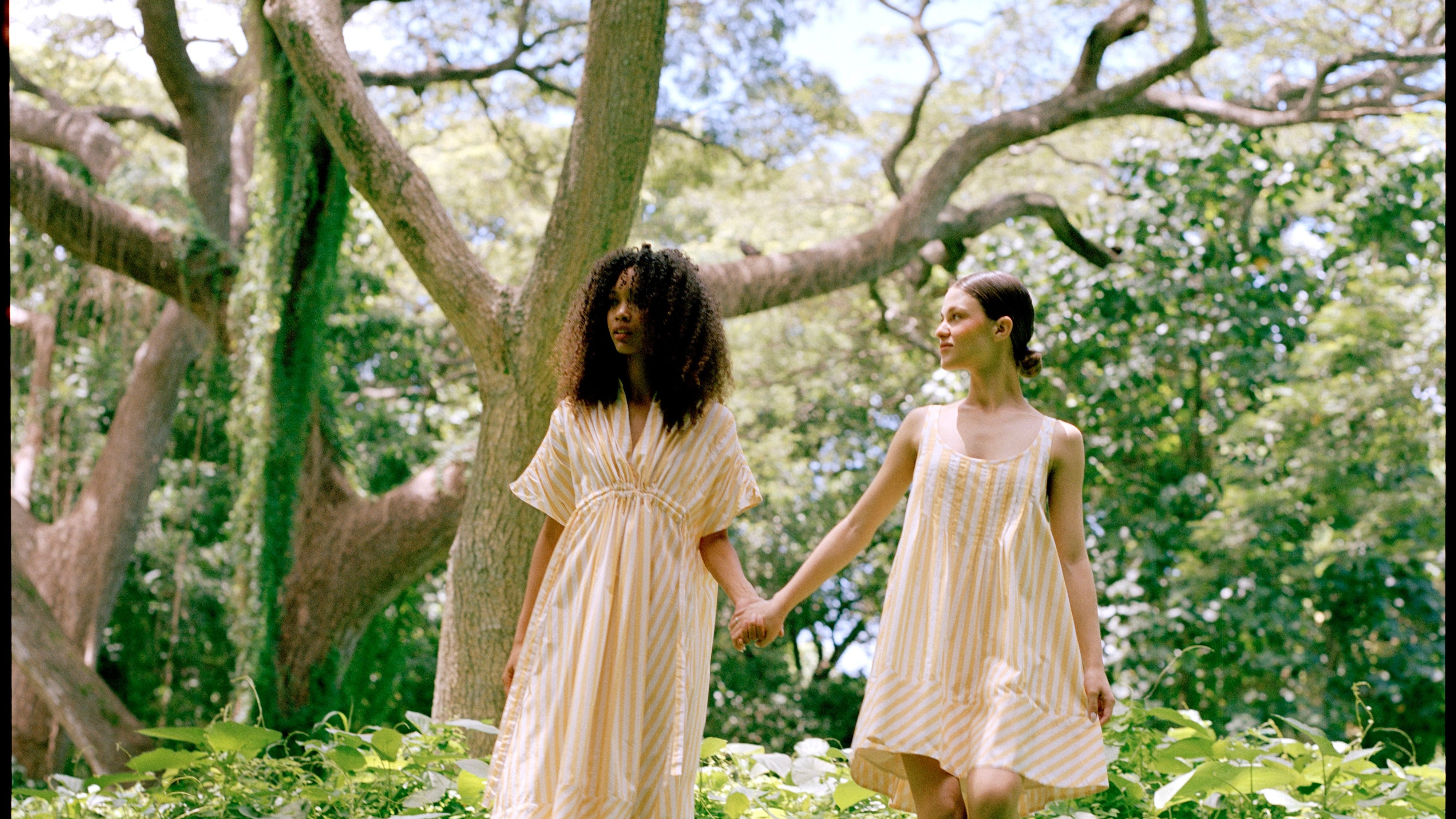 Two women in light dresses walking hand-in-hand through a lush green forest.