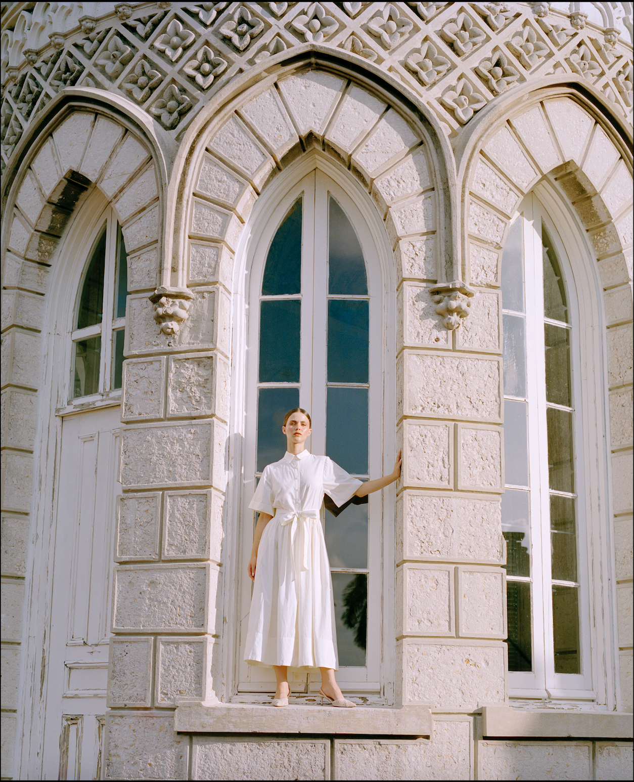 Woman in a white dress standing in front of a large, ornate window with stone architecture.